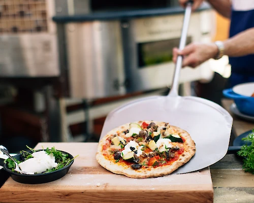A person is using a pizza peel to place a freshly baked pizza on a wooden surface, surrounded by ingredients and greenery.