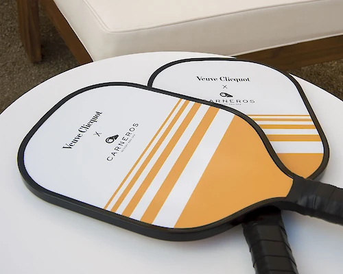 Two pickleball paddles with a white and orange design on a round white table, with a cushioned bench in the background.