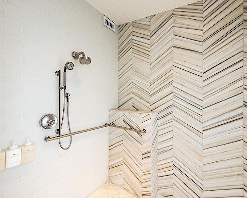 The image shows a modern bathroom shower area with a wall-mounted handheld showerhead, grab bars, soap dispensers, and stylish chevron-patterned tiles.