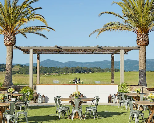 An outdoor event setup features tables and chairs under palm trees with a bar in the background, all against a scenic backdrop of rolling hills.