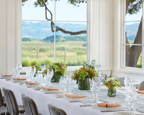 A dining setup with a long table is elegantly decorated with flowers and table settings in front of large windows showcasing a scenic countryside view.