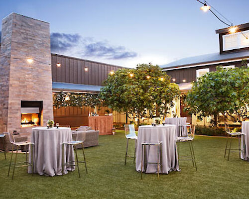 An outdoor area with tables draped in linens, chairs, a fireplace, and trees, set for an event, with string lights creating a cozy ambiance.
