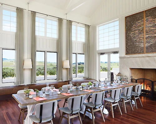 A spacious dining room with a long table, modern chairs, large windows, a fireplace, and a scenic outdoor view. The setting appears elegant and inviting.