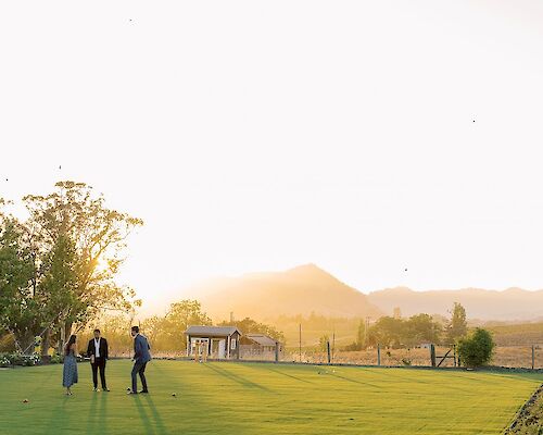 Three people stand on a lush, green field with mountains and a bright, setting sun in the background, enjoying a peaceful, picturesque scene.