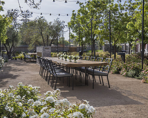 Outdoor dining area with a long table, surrounded by chairs and decorated with flowers, set in a garden with string lights and lush greenery.