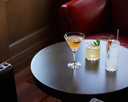 Three cocktails on a small round table: a martini, a yellow drink with mint, and a tall clear drink with a garnish, by red leather seating.