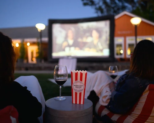 People watching an outdoor movie on a large screen, with wine glasses and popcorn in the foreground.