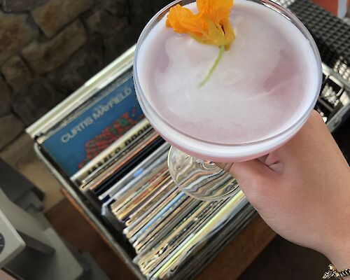 A hand holds a pink cocktail garnished with an orange flower, with a collection of vinyl records visible in the background.